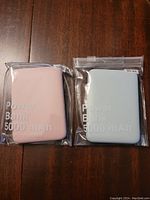 Front side of two small 5000mAh power banks, one pink and one light blue, sealed in plastic.