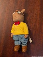 Front view of the vintage Arthur Small felt toy doll wearing yellow sweater, red bow tie, blue jeans, and brown shoes on a wooden surface.