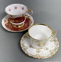 Both vintage porcelain teacups and matching saucers displayed together, showing one with deep red and gold floral design and the other with white gold-accented floral patterns.