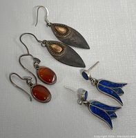 Three pairs of vintage 925 silver earrings displayed on light textured fabric. Includes large teardrop dangles with patina and copper insert, smaller oval red/orange stone dangles, and blue lapis floral stud earrings.