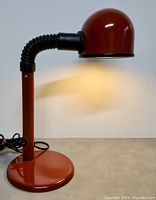 Side view of red Alda desk lamp switched on, showing dome-shaped shade and flexible gooseneck arm.