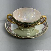 Front view of antique Limoges double handle teacup resting on matching saucer showing hand painted floral and gold details and iridescent finish.