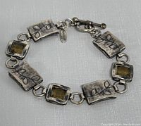 Close-up image showing the engraved rectangular links and yellow stones, along with toggle clasp and attached small metal tag.