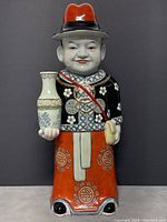 Front view of the Chinese porcelain statue showing the figure holding a vase and wearing traditional clothing with a red hat.