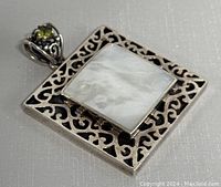 Front view of the square sterling silver pendant with central mother of pearl and decorative cutout design in the frame, bail with peridot accent visible