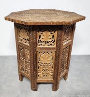 Side view of the fully assembled octagonal hand carved rosewood folding side table showing detailed floral and leaf carvings on vertical panels and scalloped tabletop edge.