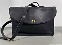 Front view of the black leather Coach briefcase featuring the flap with gold twist-lock closure and leather shoulder strap draped over front.