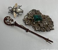 Three vintage clip and brooch jewelry pieces on white background including a jewel-toned brooch, red rhinestone sweater clip, and bee fur clip.