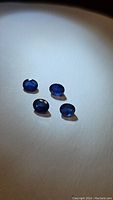 Four natural oval cut deep blue sapphires arranged on a light surface under spot lighting; stones measured approx. 5x4mm each, showing natural inclusions.
