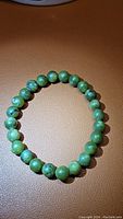 Green jade bead bracelet with round 6mm beads lying on a brown surface, showing natural color variation and polished finish.