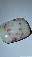 Close-up image of a rectangular cushion cabochon gemstone with a white base color and red-pink cinnabar inclusions embedded inside.