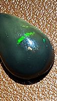 Close-up image of a pear-shaped natural black opal cabochon showing green play of color on dark background.