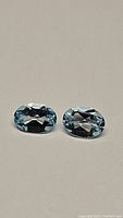 Pair of oval cut sky blue topaz gemstones lying side by side on a plain light background, showing their clarity and color.
