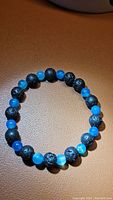 Top view image showing the natural gemstone bracelet arranged in a circle on a tan surface, highlighting the alternating black lavastone and blue agate beads.