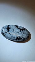 Photo of an 18.85ct natural oval cabochon made of snowflake obsidian gemstone showing detailed snowflake-like patterns in black and gray.