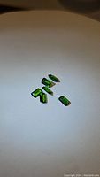 Photo showing six emerald cut bright green chrome diopside gemstones arranged on a beige surface.