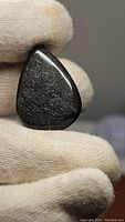 Front view of pear-shaped silver sheen obsidian cabochon held with a gloved hand showing the reflective metallic sheen on the polished surface.