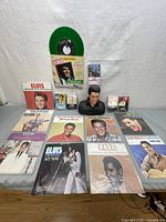 Displays 13 Elvis Presley records arranged on the floor with one green record in its sleeve upright, showing a variety of album covers including recent releases and classics.
