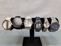 Front view of six wrist watches on black display stand, showing various dial colors and band types