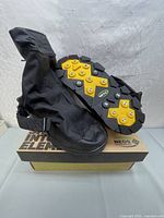 Pair of NEOS overshoe boots positioned on top of NEOS branded shoebox. One boot shows the sole with yellow cleats and metal studs.