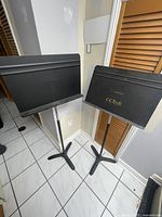 Two black Manhasset music stands positioned side by side on tiled floor showing overall condition and size.