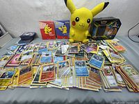 Wide shot displaying a large pile of assorted Pokémon cards with two binders, Pikachu plush, and collectible tin in background.