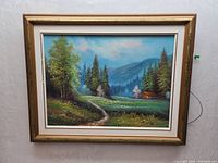 Framed oil painting showing a forest scene with trees, a cabin, a stream, and mountains in the background.