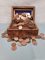 Wooden treasure chest overflowing with copper pennies, blue felt lining visible inside lid, pennies scattered around chest on flat surface