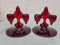 Pair of red glass 2-light candle holders showing front view with fleur-de-lis style backplate and round base.