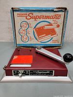 Front angled view showing the cigarette rolling machine and original box behind it, highlighting the crank handle and the bright orange label on the top.