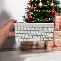 Apple wireless Bluetooth keyboard with white keys and aluminum body shown held in hand in front of Christmas decorations.