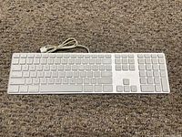 Top down view of Apple white wired keyboard on carpet showing full keyboard including numeric keypad and USB cable coiled at top left.