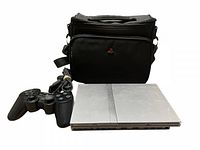 Silver Sony PlayStation 2 slim console, controller, and carry bag shown together with cables, console top view revealing wear but intact branding.