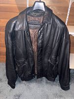 Front view of the black leather jacket on a hanger, showing full length zipper, collar, flap pockets, and elastic waist and cuffs