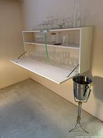 Shelf with multiple types of clear drinking glasses arranged including stemmed wine glasses, flutes, and tumblers, plus a stainless steel ice bucket on a tripod stand next to the shelf.