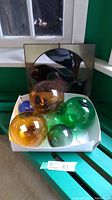 Set of six glass gazing balls of amber, green, blue colors in a white box on a green chair near a window