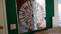 Photo showing the square mirror hanging on a green wall with a detailed circular geometric pattern in white on the mirrored surface, reflecting the room and hallway.