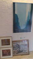 Four framed artworks displayed on floor and wall: an abstract blue-gray print on wall and three smaller landscape style works on floor (two vertical, one horizontal).