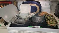 Overview of assorted kitchen items on stove including white casseroles, small ceramic dishes, clear serving bowls, blue woven place mats, and decorative hotplates.