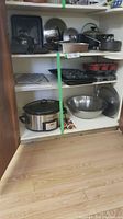 Full view of kitchen shelving holding multiple pots, pans, a slow cooker, metal mixing bowl, baking pans and muffin tins.