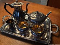 Full set of vintage silver plated tea service on tray showing coffee pot, teapot, creamer, sugar bowl, small cup, and tray with ornate handles.