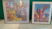 Pair of gold-framed scenic prints depicting autumn forest scenes with fall foliage in warm colors, displayed side-by-side on carpeted floor.