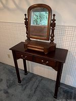 Vintage wooden vanity mirror on a wooden side table with two drawers, carpeted floor and wallpaper background.