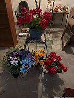 Artificial red flowers in blue plastic pot on metal plant stand, other flower pots and vase on floor around it