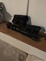 Photo of the tripod, carrying bag, and protective case on a wooden shelf next to a white wall.
