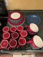 Photo showing a stack of red and white rimmed dinner plates, blue side plates, red mugs, and red and white cups with blue trim on a countertop.