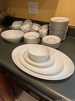 Full set of white china dinnerware laid out on kitchen counter showing plates, bowls, cups, and serving dishes.