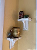 Two white wall shelves holding three decorative boxes and one bird figurine against yellow wall.
