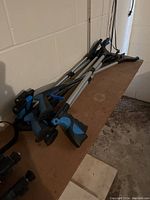 Seven grabber reaching tools with blue and black ergonomic handles and silver shafts are shown lying on a wooden surface against a white brick wall.