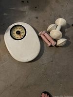 White mechanical bathroom scale with a dial showing weight, placed on floor next to three pairs of dumbbells (white and pink).
